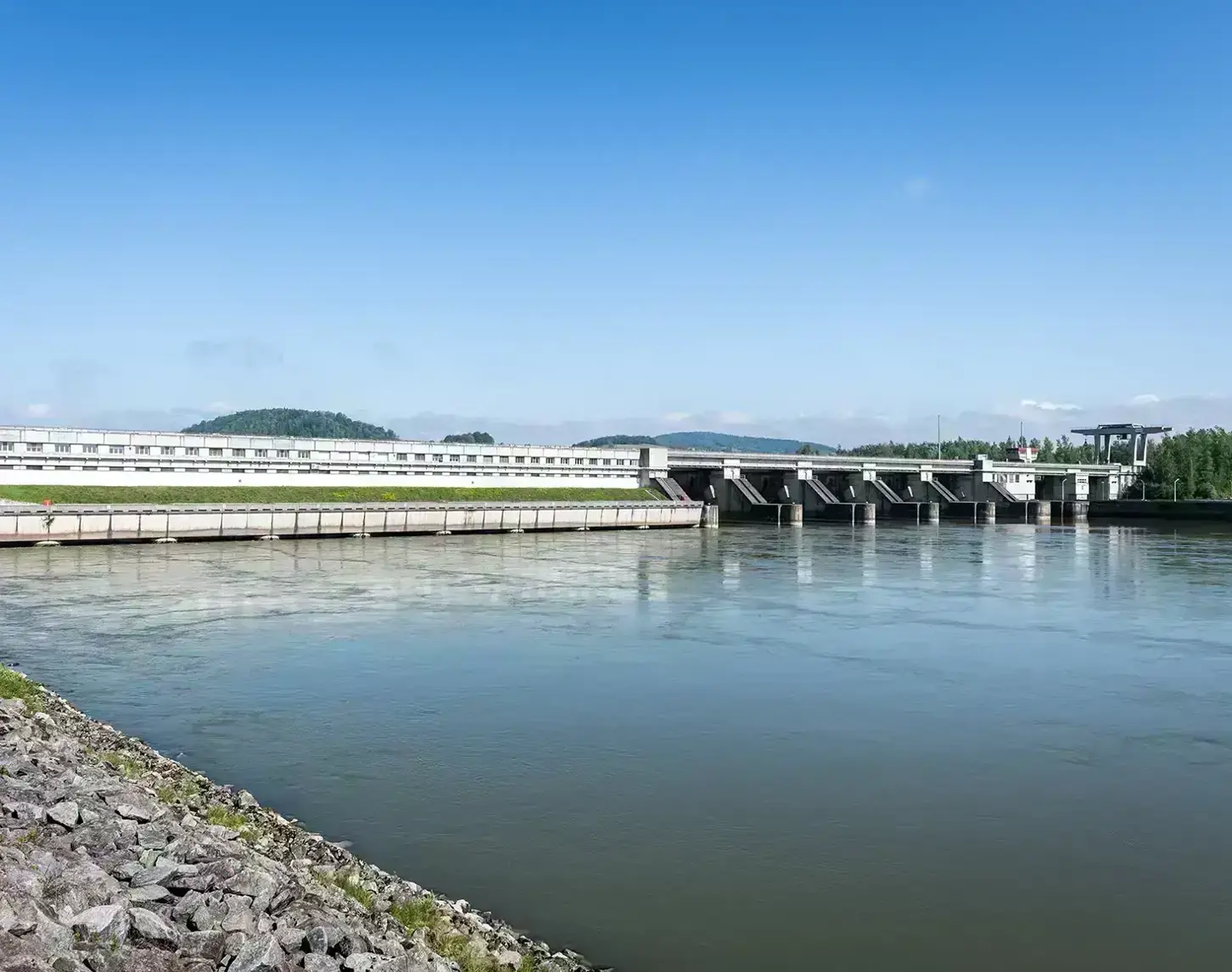 Blick auf das Kraftwerk Abwinden Asten. Links sieht man einen Teil der Uferbegrenzung. Der blaue Himmel spiegelt sich in der Donau.