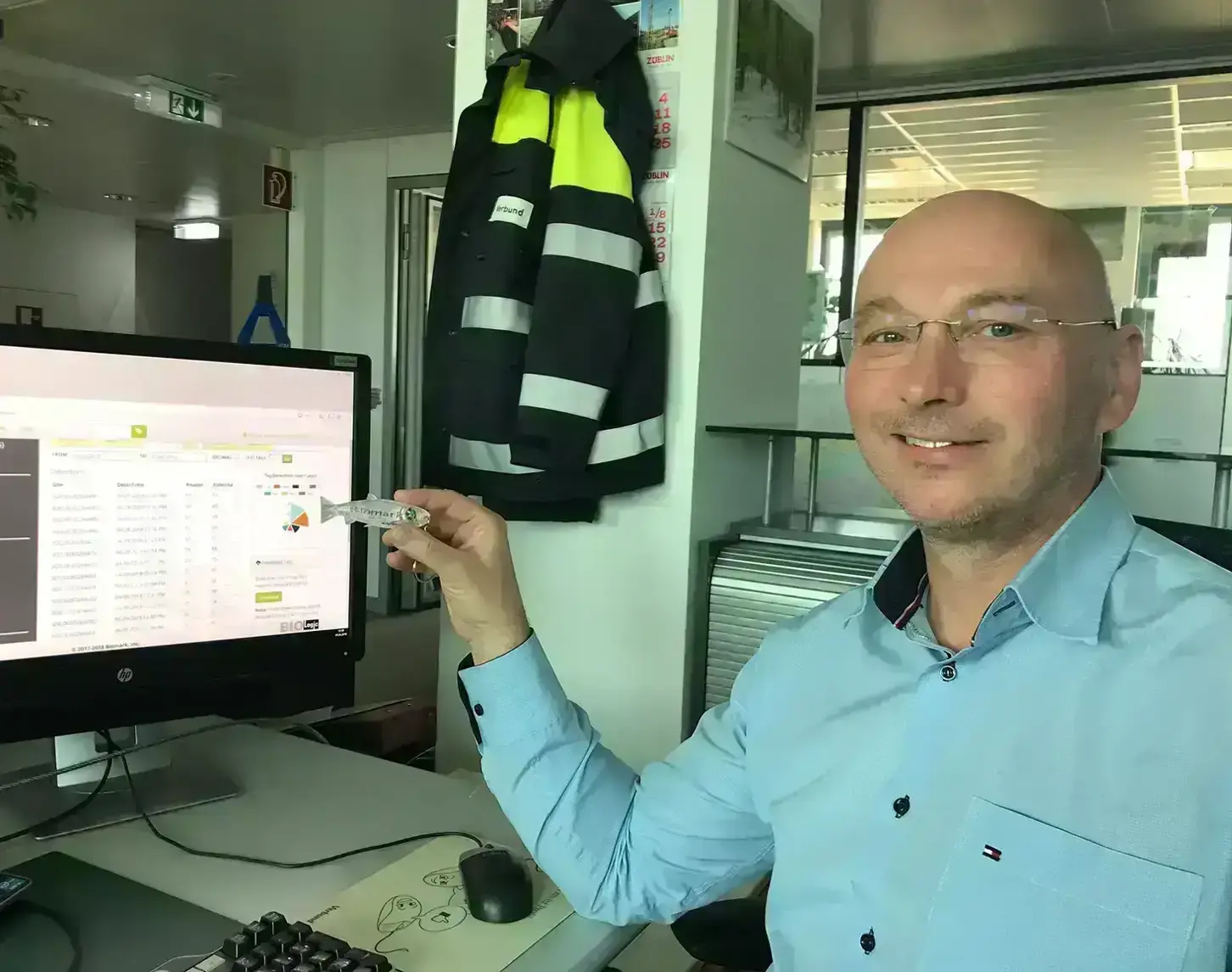 Walter Reckendorfer, an employee at VERBUND, shows our editor how fish monitoring works. They are sitting in front of a computer and Walter shows a small fish to the camera.